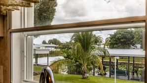 Vista de dentro de um quarto pela janela, mostrando uma área gramada com uma palmeira, um balanço e um espaço de estar coberto.