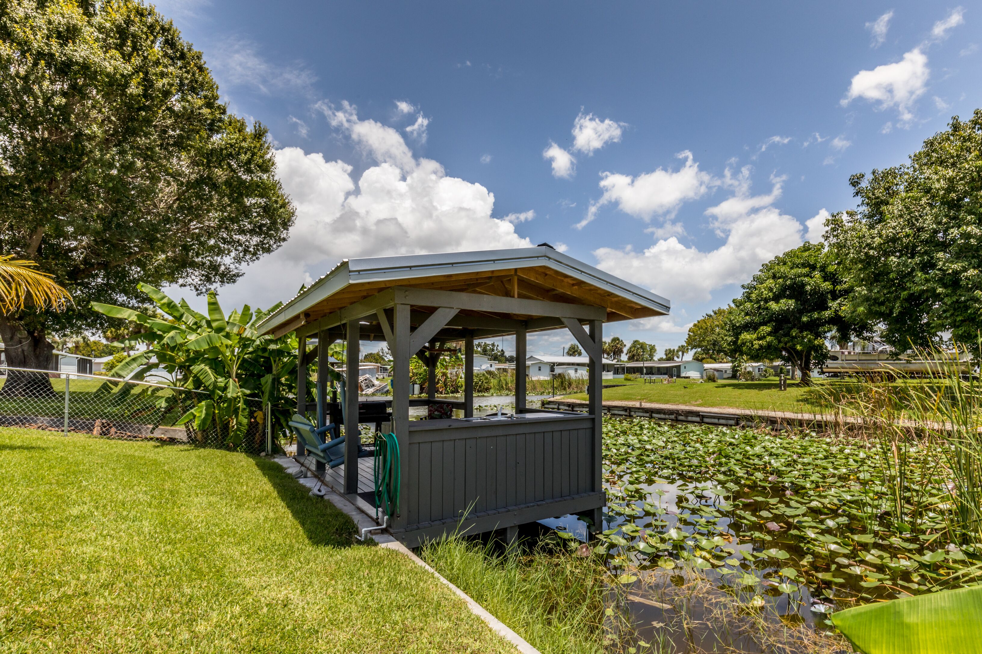 A gazebo with a wooden roof and walls, surrounded by a pond with lily pads and a grassy area.