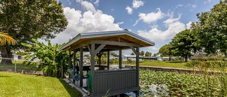 A gazebo with a wooden roof and walls, surrounded by a pond with lily pads and a grassy area.