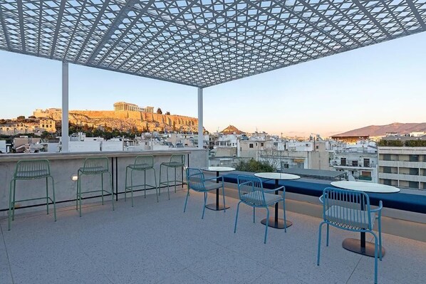 A rooftop terrace with outdoor seating, a view of ancient ruins, and a modern metal roof structure. - Acro Urban Suites (Athens)