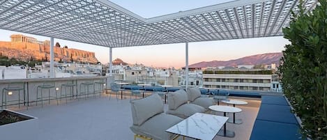 An outdoor seating area with a view of the Acropolis, featuring modern furniture and a pergola structure.