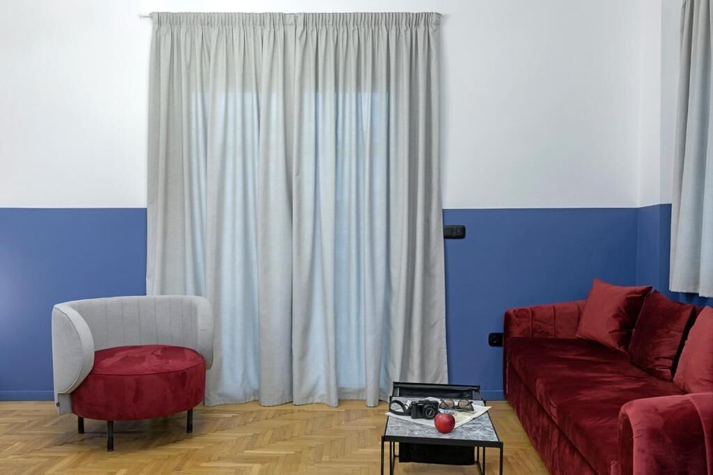 A room with a blue and white wall, a grey curtain, a red sofa, a grey armchair, and a small table with a camera and an apple.