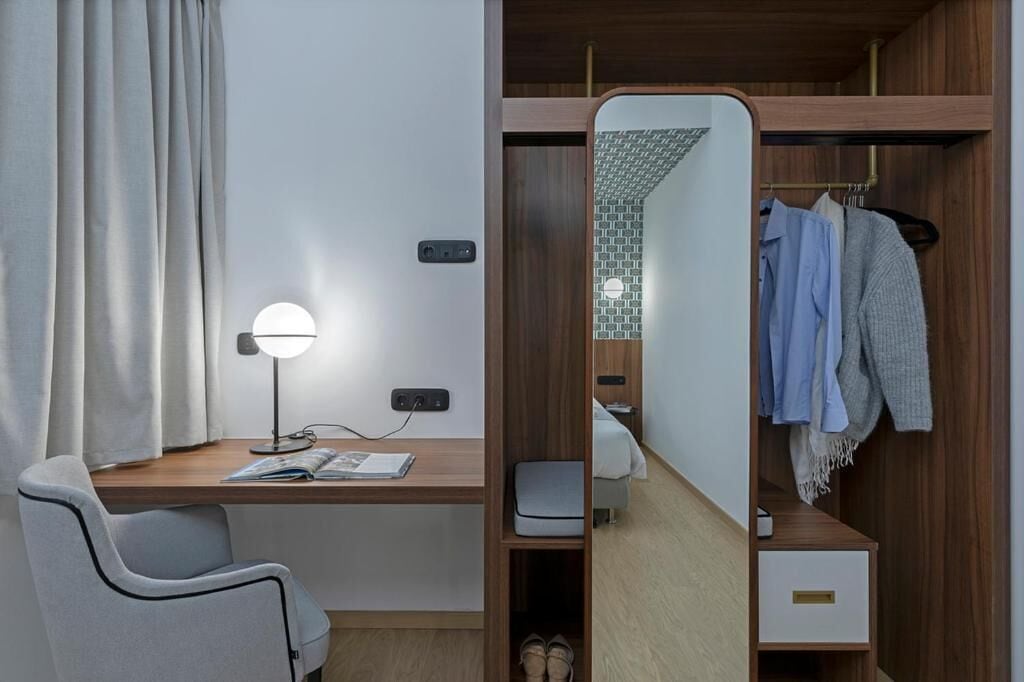 Una camera compatta e moderna con una scrivania in legno, una sedia, un comodino, uno specchio e un armadio con abiti appesi.