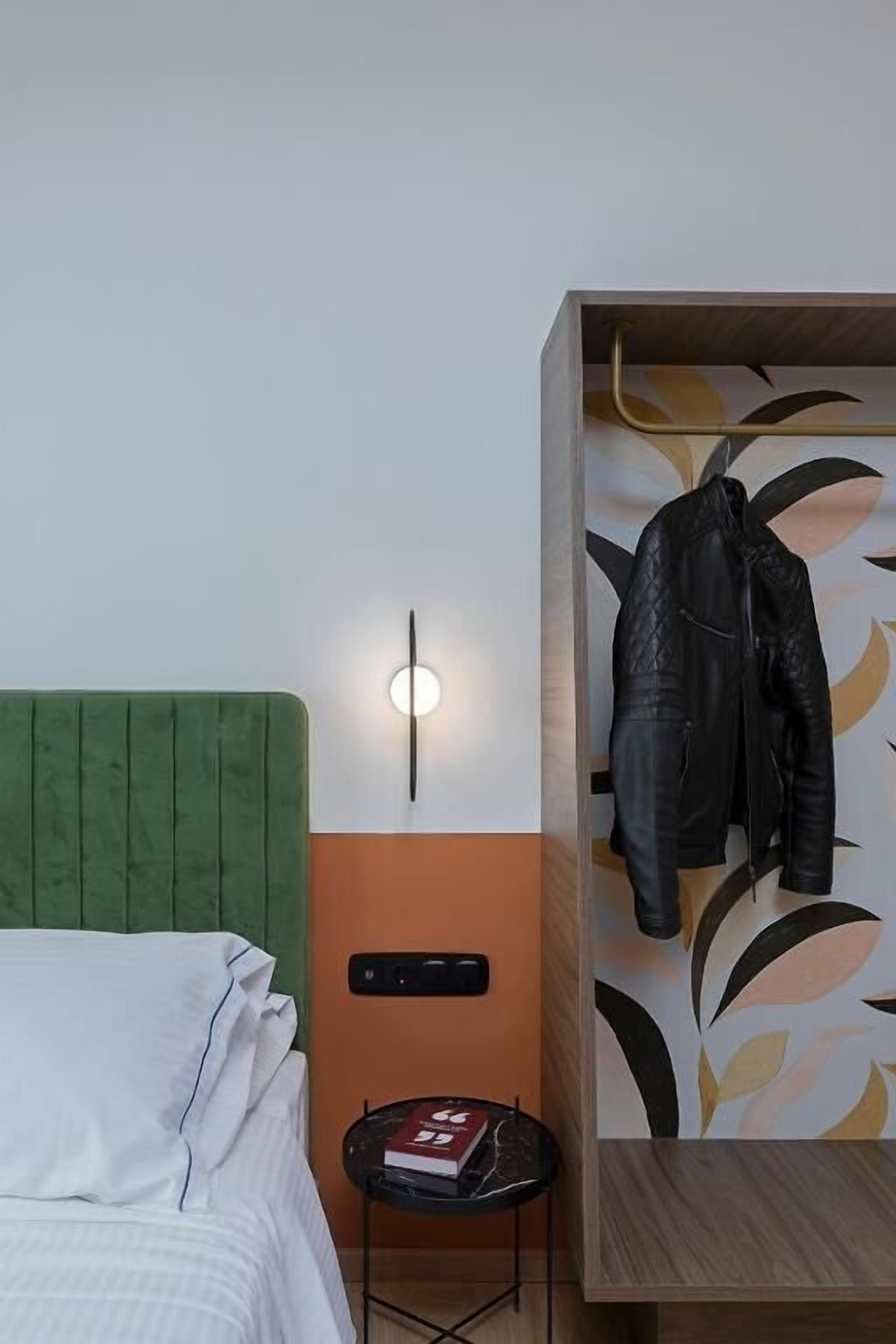 A room with a green upholstered headboard, a small table with a book, a wall-mounted light, and a wooden cabinet with a jacket hanging on it.
