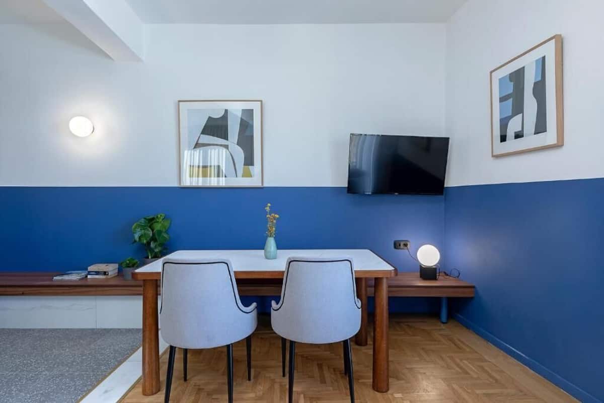 A room with a wooden dining table, two chairs, a TV mounted on the wall, and framed artwork.