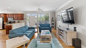 A spacious living room with a large TV, two sofas, a coffee table, and a kitchen area with wooden cabinets and a microwave.