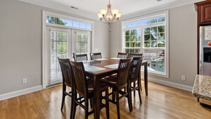 A dining room with a wooden table and chairs, large windows, and a chandelier.