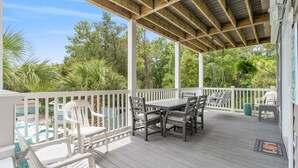 A covered outdoor patio with a wooden table and chairs, surrounded by white railings and lush greenery.