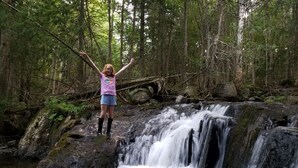 Una persona de pie sobre una roca con los brazos levantados en un área boscosa con una cascada.