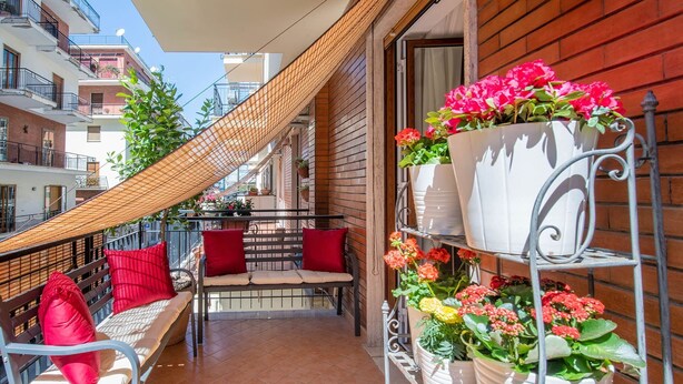 Ein Balkon mit Korbstuhl, rotem Kissen, kleinem Tisch und Topfpflanzen.