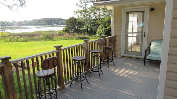 A wooden deck with four bar stools and a single chair, overlooking a grassy area and a body of water.