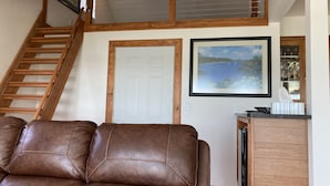 Een woonkamer met een bruine leren bank, een houten trap, een witte deur en een lijst met een schilderij van een kustlandschap.