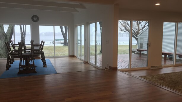 Een ruime kamer met houten vloer, een eettafel met stoelen, en grote ramen met uitzicht op een terras en een gebouw aan de overkant van het water.