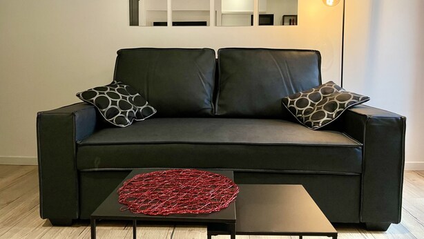 Ein modernes Wohnzimmer mit einer schwarzen Ledersofa, zwei gemusterten Kissen, einer roten abstrakten Skulptur und einem Stadtbild an der Wand.
