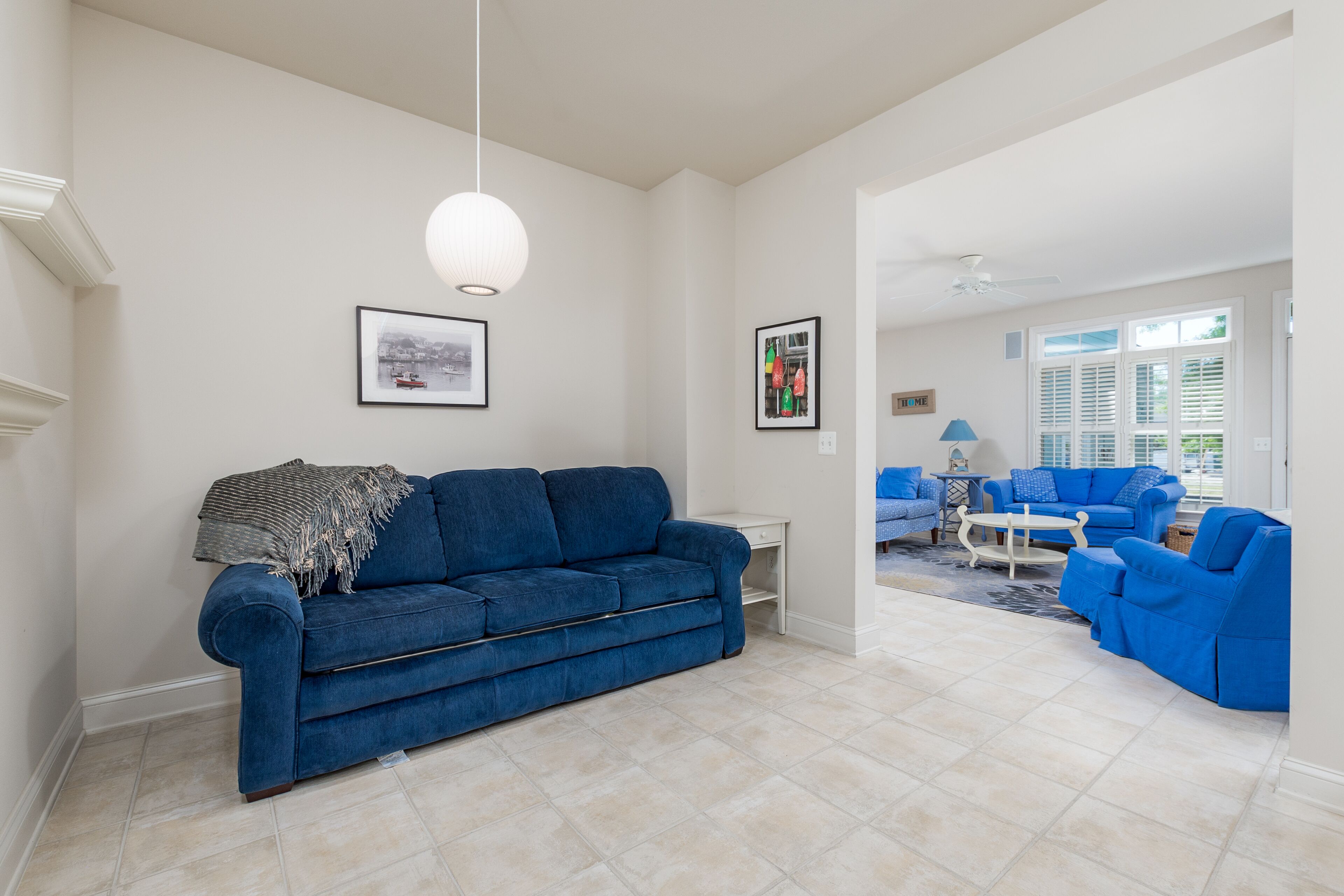 Un salon avec un canapé bleu, une table basse blanche et un fauteuil bleu. Les murs sont ornés de tableaux encadrés et un ventilateur de plafond est installé au centre.