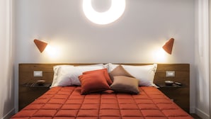 Ein ordentlich bezogenes Bett mit einer roten Steude, zwei Nachttische mit Schalen und eine runde Leuchte darüber.