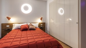 Ein Schlafzimmer mit roter gesteppter Bettdecke, zwei Nachttischen mit Lampen, einem Kleiderschrank und runden Wandleuchten.