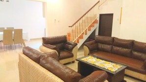 A living room with a brown leather sofa, a coffee table with a floral table runner, and a staircase with wooden railings. - Fams Homestay by FH Stay (Baturetno)