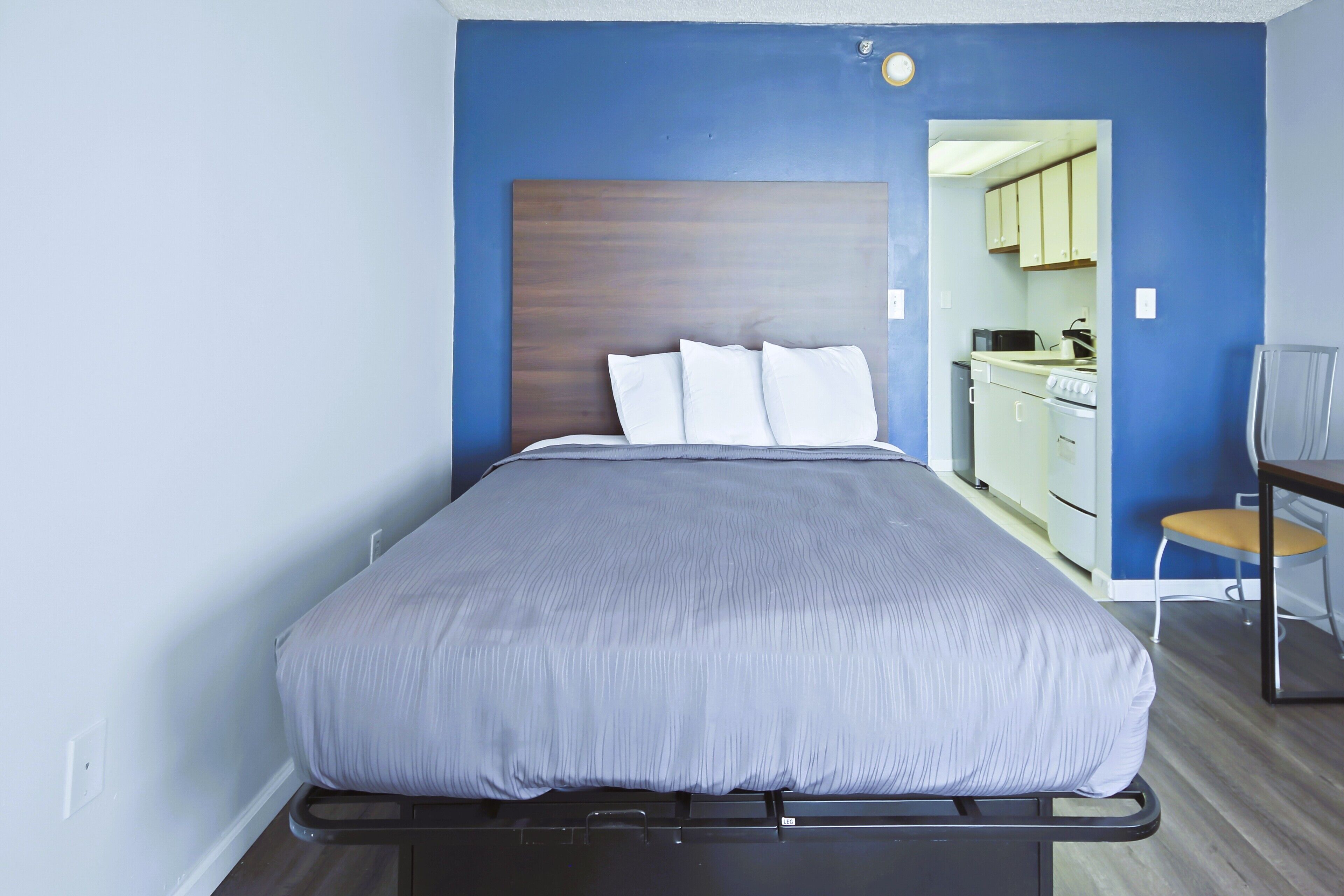 A bedroom with a bed, a wooden headboard, a chair, and a view into a kitchen area.