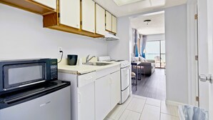 Cocina compacta con microondas, pileta y estufa. Se ve un comedor con sofá y mesa a través de un umbral abierto.