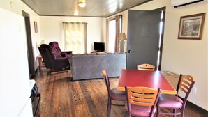 Una habitación con piso de madera, una mesa de comedor con cuatro sillas, un sofá, un televisor y una puerta.