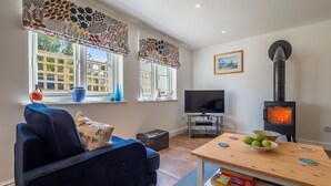 A living room with a dark blue sofa, a wooden table with a bowl of apples, a television, and a black wood-burning stove. - Yew Tree Cottage is a modern eco-cottage (Westleton)