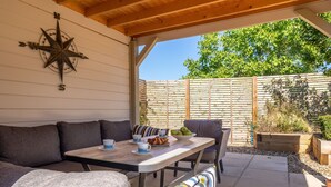 A covered patio with a wooden table, grey sofa, and a star compass wall decoration. - Yew Tree Cottage is a modern eco-cottage (Westleton)