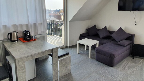 Un salon moderne avec un canapé gris, une petite table blanche et un coin cuisine équipé d’une bouilloire et d’une machine à café.