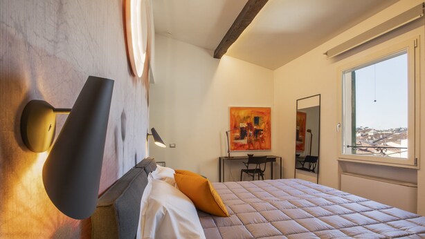 Un dormitorio con cama, mesita de noche, lámpara, un cuadro, ventana y vista de la ciudad.