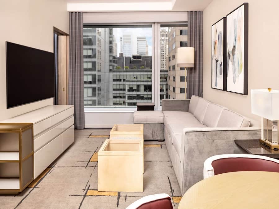 A modern hotel room with a large window, a flat-screen TV, a sofa, a dining table, and a side table with a lamp.