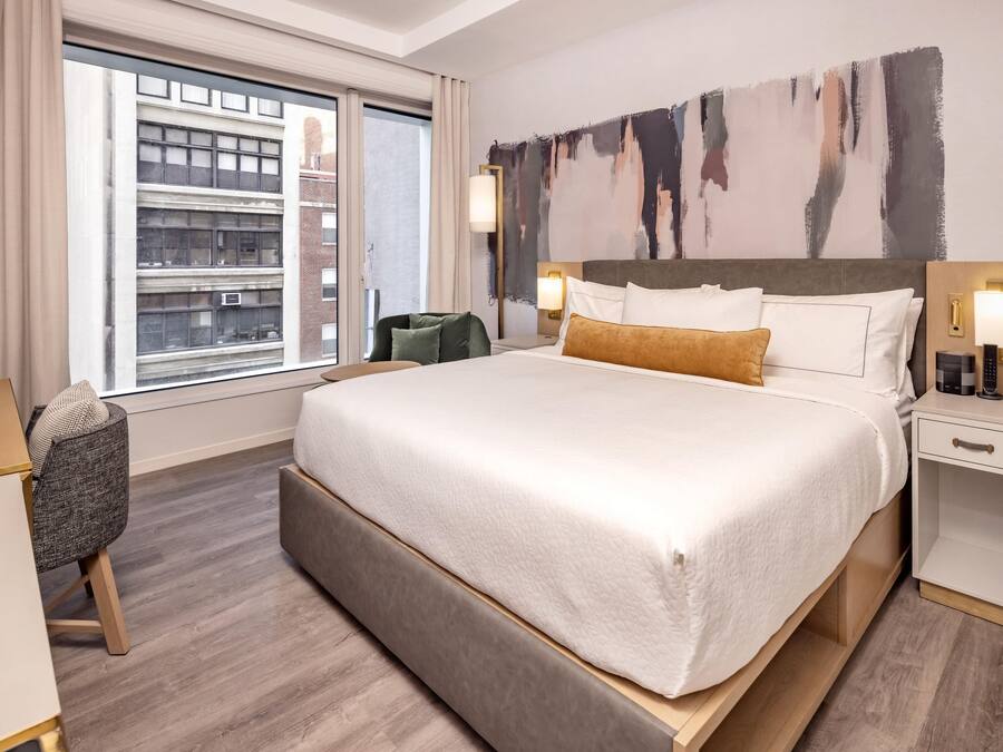 A hotel room with a large bed, a desk, a chair, and a view of the city through the window.