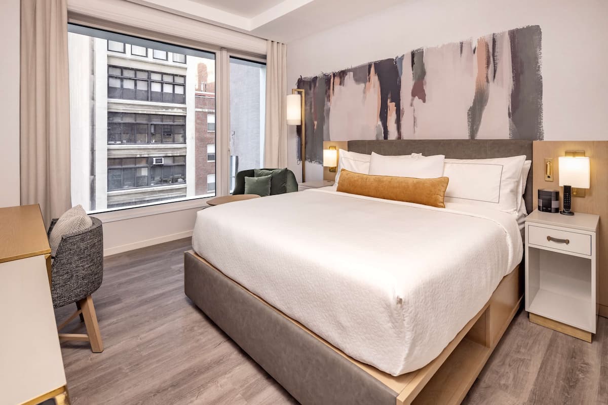 A hotel room with a large bed, a desk, a chair, and a view of the city through a window.