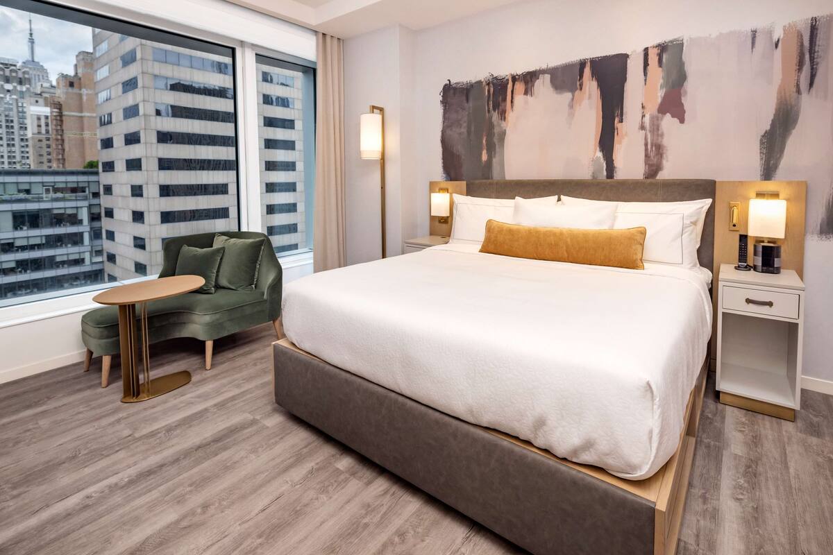 A modern hotel room with a large bed, a green armchair, a small table, and a city view through large windows.
