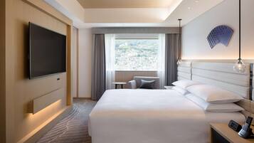 A hotel room with a large bed, a flat-screen TV mounted on the wall, a small table, a chair, and a view of the city through the window.