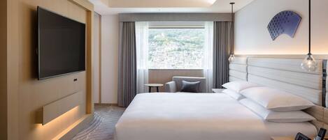 Habitación de hotel con una cama grande, un televisor de pantalla plana montado en la pared, una mesita, una silla y vistas de la ciudad desde la ventana.