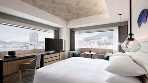 Una habitación de hotel moderna con una cama grande, un escritorio con televisor de pantalla plana, una mesita pequeña y vistas a un paisaje urbano a través de amplios ventanales.