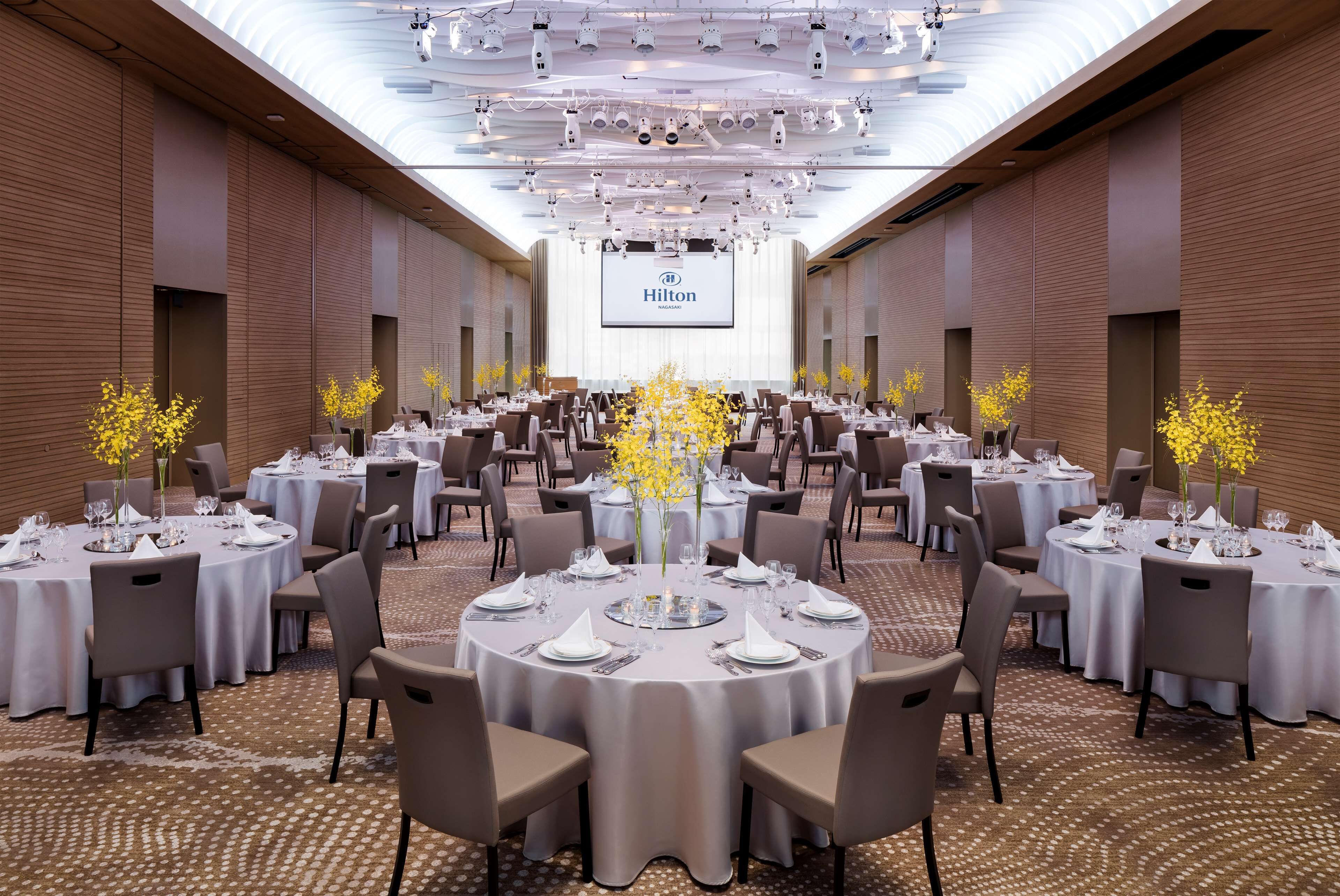 Una amplia sala de conferencias bien iluminada, con mesas redondas dispuestas para comidas, una gran pantalla mostrando el logotipo de Hilton, y un diseño de techo moderno.
