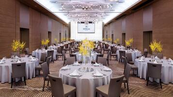 Una amplia sala de conferencias bien iluminada, con mesas redondas dispuestas para comidas, una gran pantalla mostrando el logotipo de Hilton, y un diseño de techo moderno.