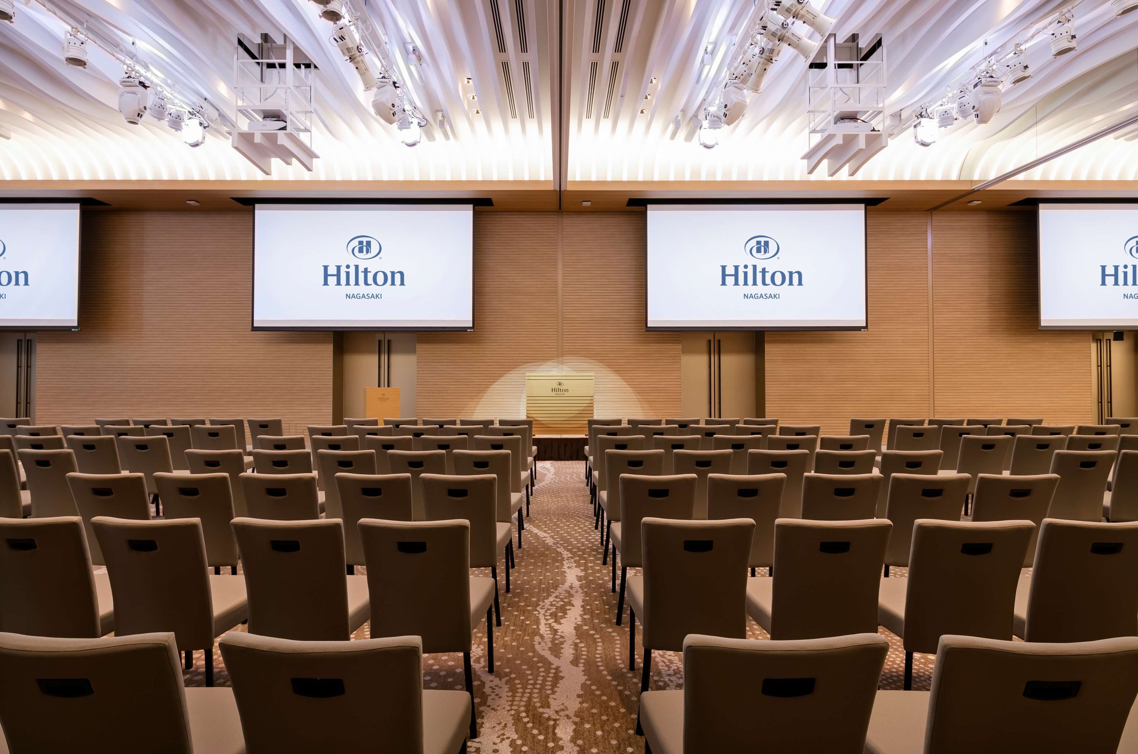 A conference room with rows of beige chairs, a carpeted floor, and a ceiling with lighting fixtures.