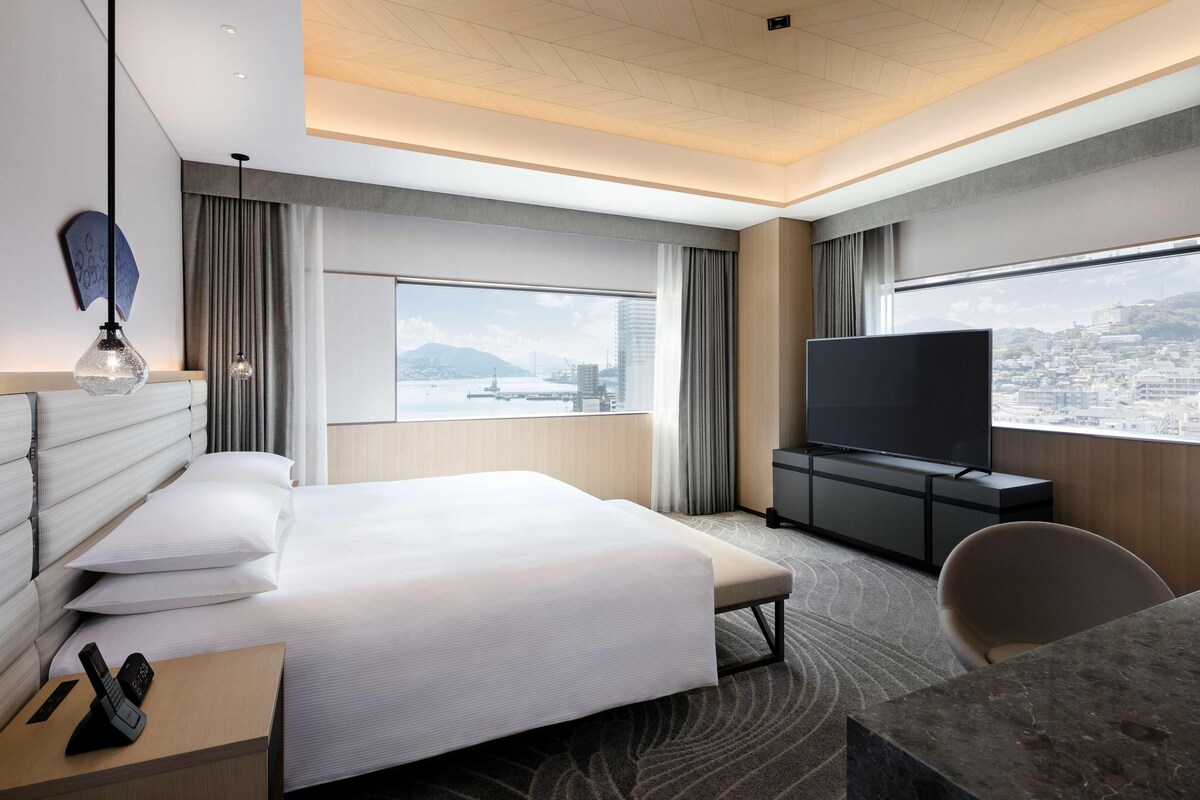 A modern hotel room with a large bed, a TV, a desk, and a view of the city.