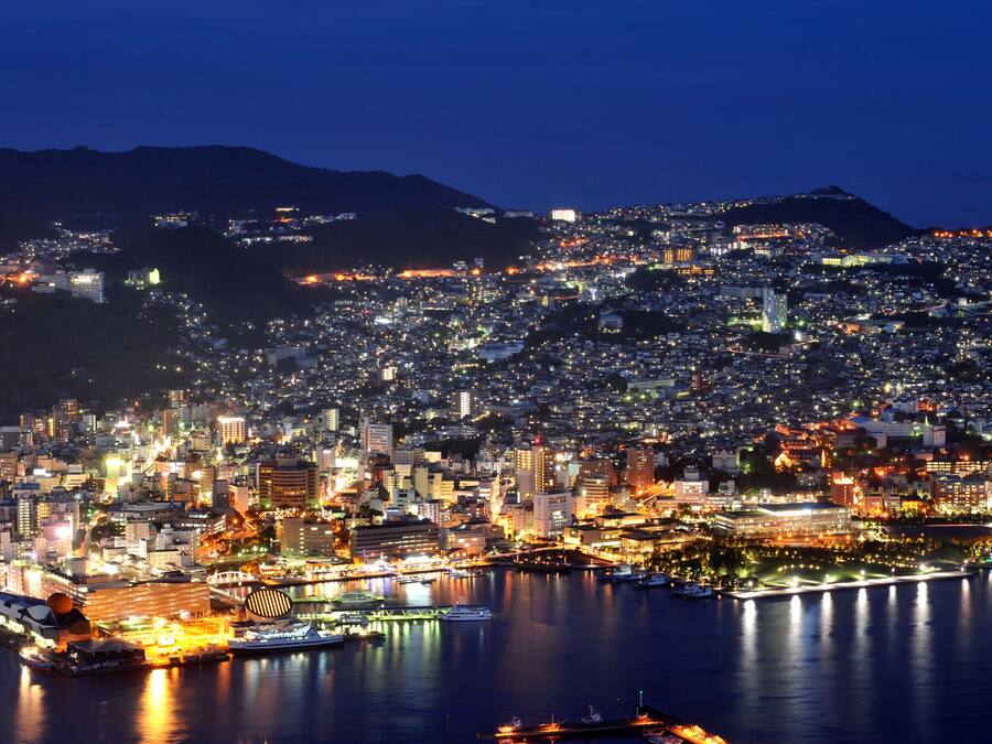 A nighttime cityscape with a densely populated urban area, a prominent waterfront, and a bridge connecting two parts of the city.