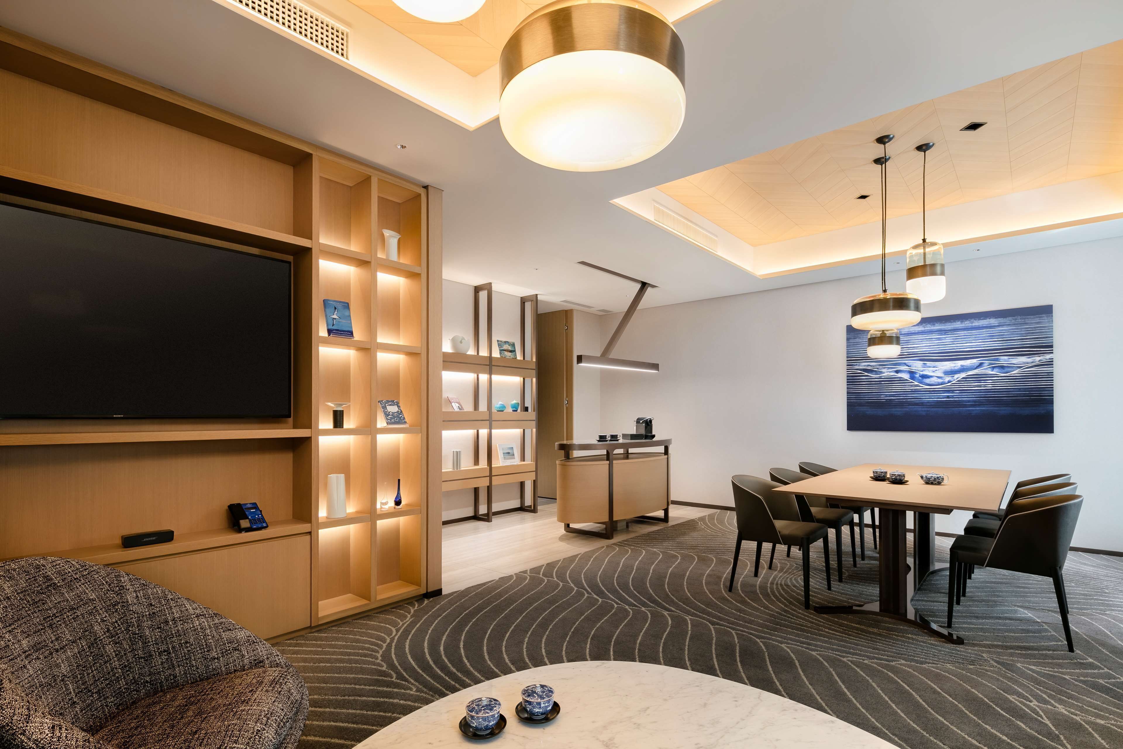 A modern hotel room with a flat-screen TV, a dining area with a table and chairs, a sofa, and a large painting on the wall.