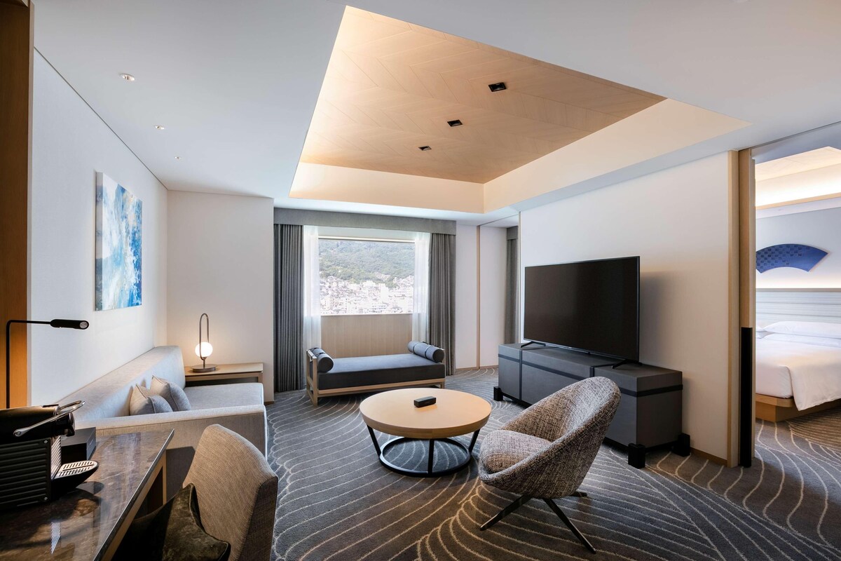 A modern hotel room with a sofa, a round coffee table, a TV, a desk with a lamp, and a bed.