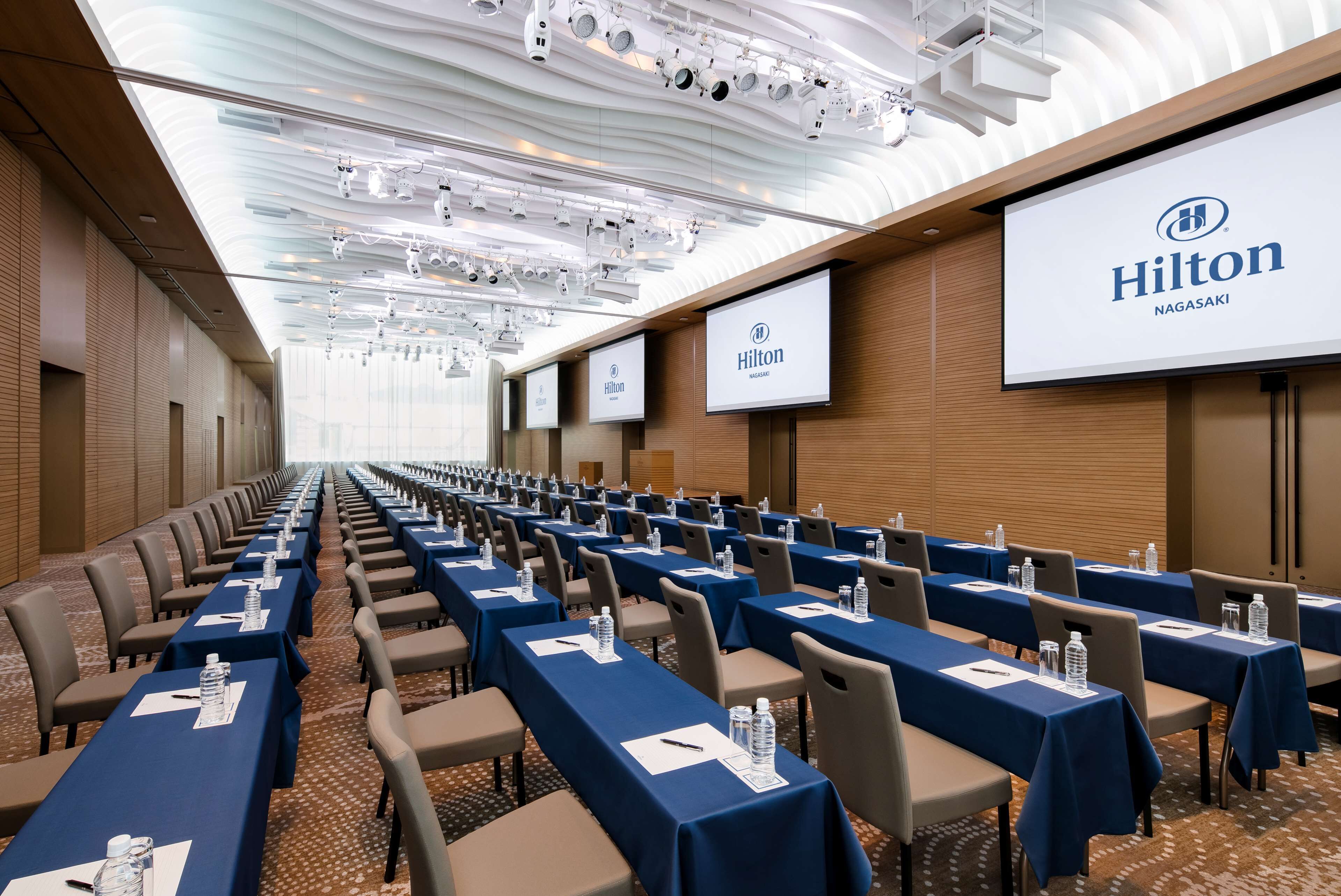 A conference room with rows of tables, each with a blue tablecloth, water bottles, and name cards. The room has a high ceiling with recessed lighting and a large screen displaying the Hilton logo.