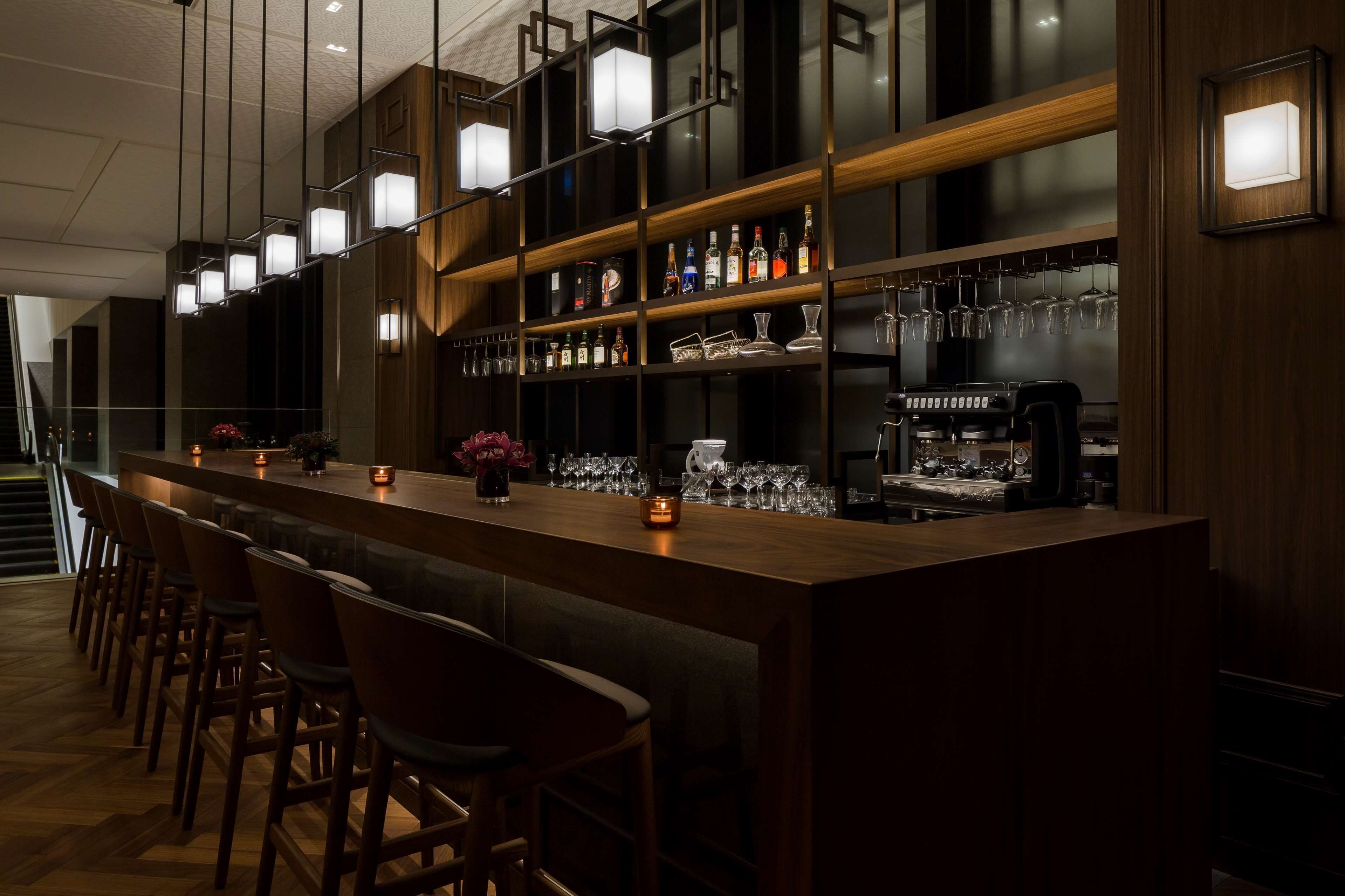 A bar area with a long counter, multiple stools, shelves with bottles and glasses, and a coffee machine.