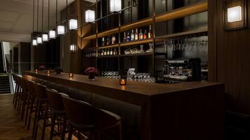 A bar area with a long counter, multiple stools, shelves with bottles and glasses, and a coffee machine.