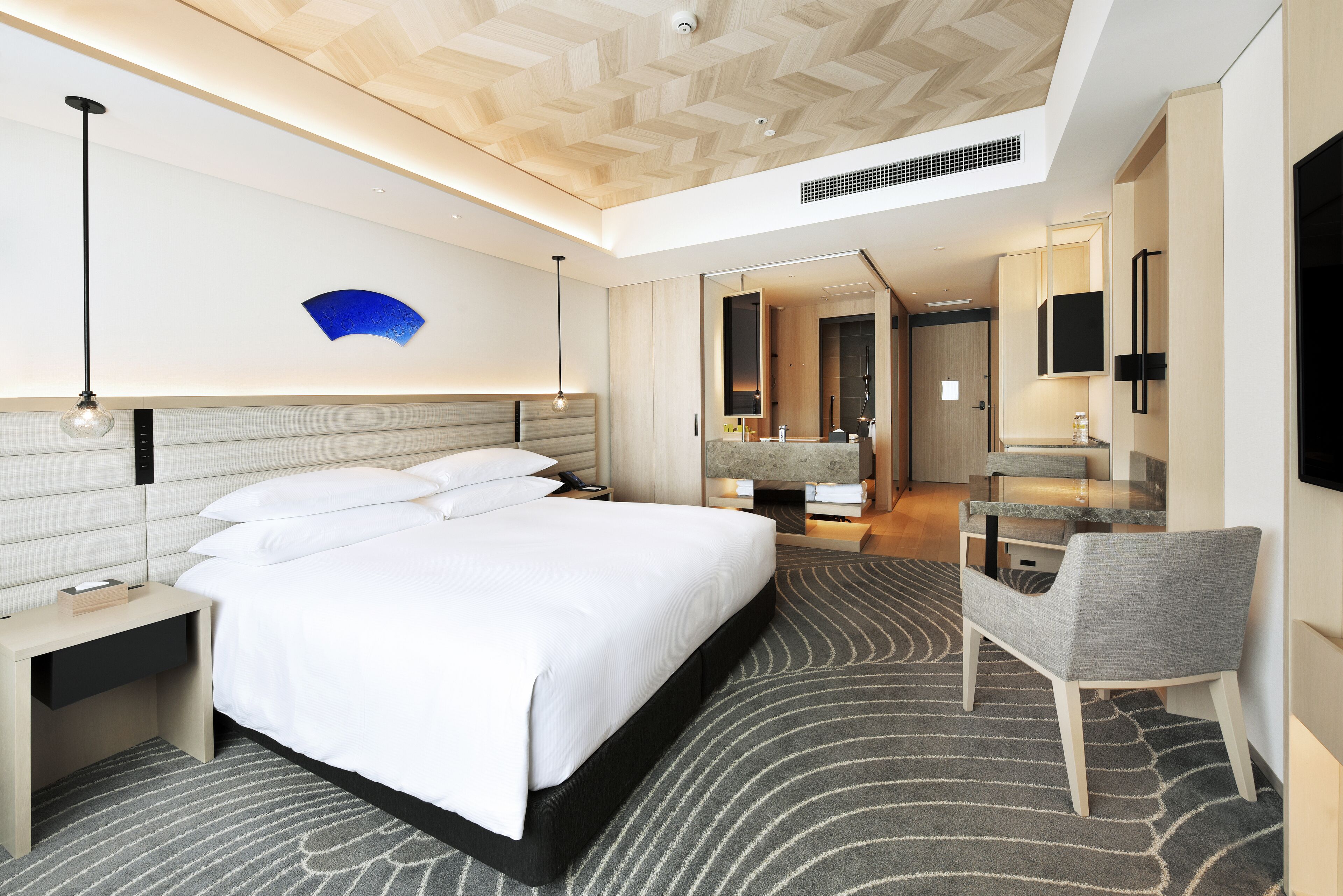 Una habitación de hotel moderna con una cama grande, mesitas de noche, una silla, un espejo y un baño visible a través de una puerta abierta.