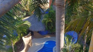 Un espace piscine agrémenté d’un toit de chaume, de palmiers et d’un élément décoratif aquatique aux eaux bleues.