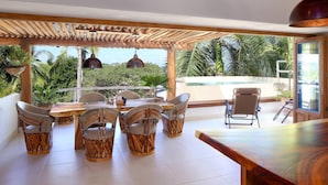 Ein großzügiger Außenbereich mit Essbereich, einer Holz-Pergola, Korbstühlen und Blick auf Palmen und einen Pool.