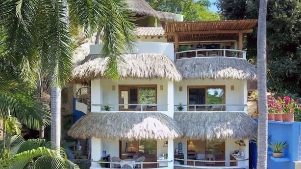 A multi-story building with a thatched roof, surrounded by lush greenery and palm trees.